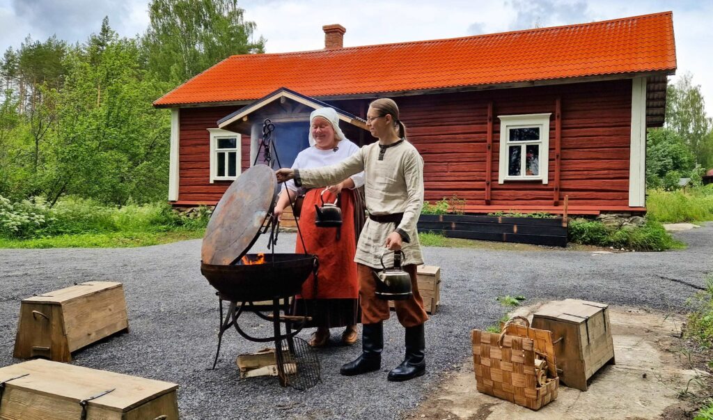 Muinaispukuiste nainen ja mies rautapadassa olevan nuotin äärellä keittämässä nokipannukahvia punaisen hirsituvan pihamaalla. 